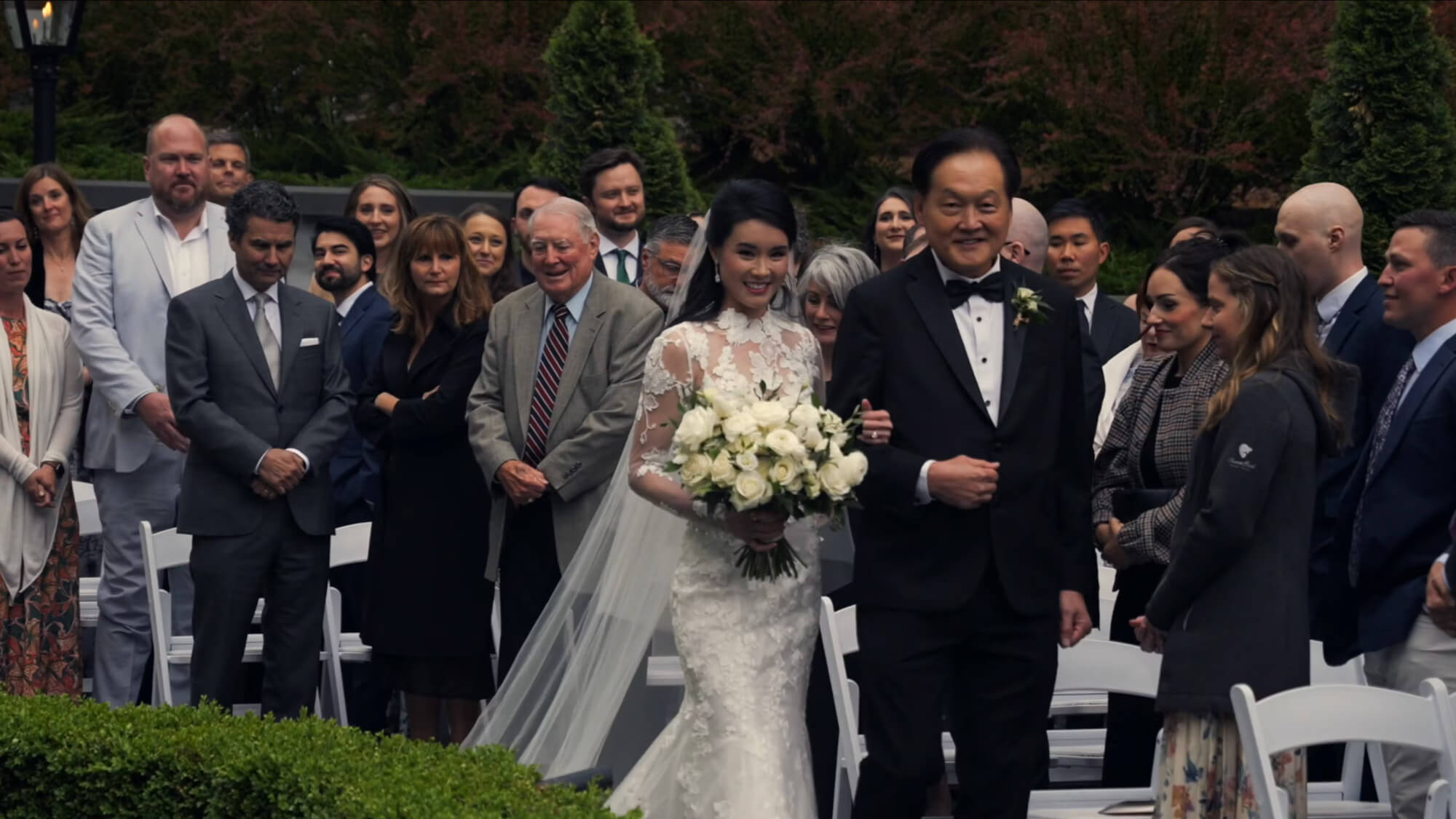 A bride in a white lace gown holding a bouquet walks down an outdoor aisle with a man in a black tuxedo. Guests on both sides stand and watch. Greenery and trees are visible in the background.