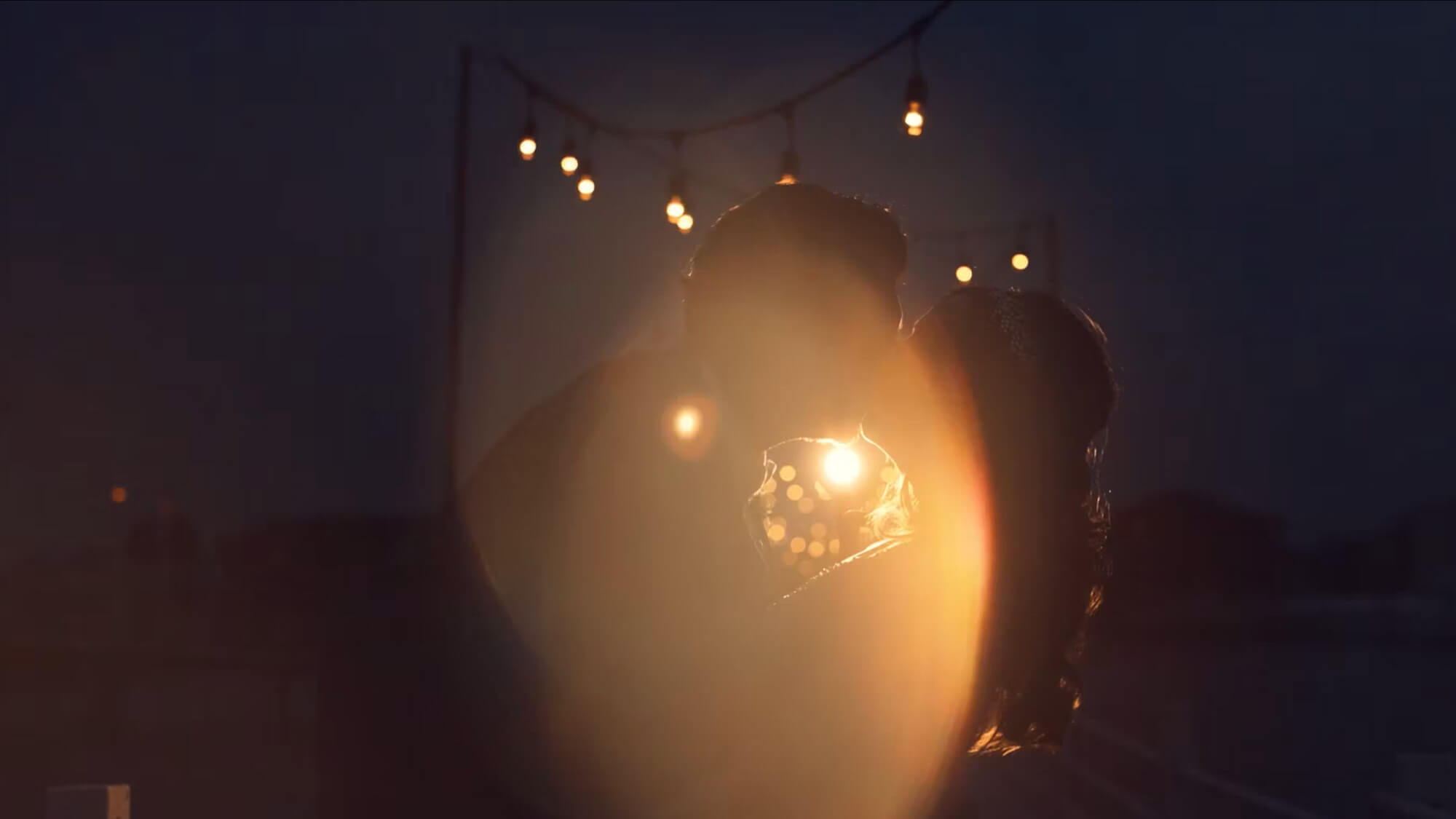 Two people are facing each other closely at night with string lights hanging in the background and a bright lens flare partially obscuring their faces.