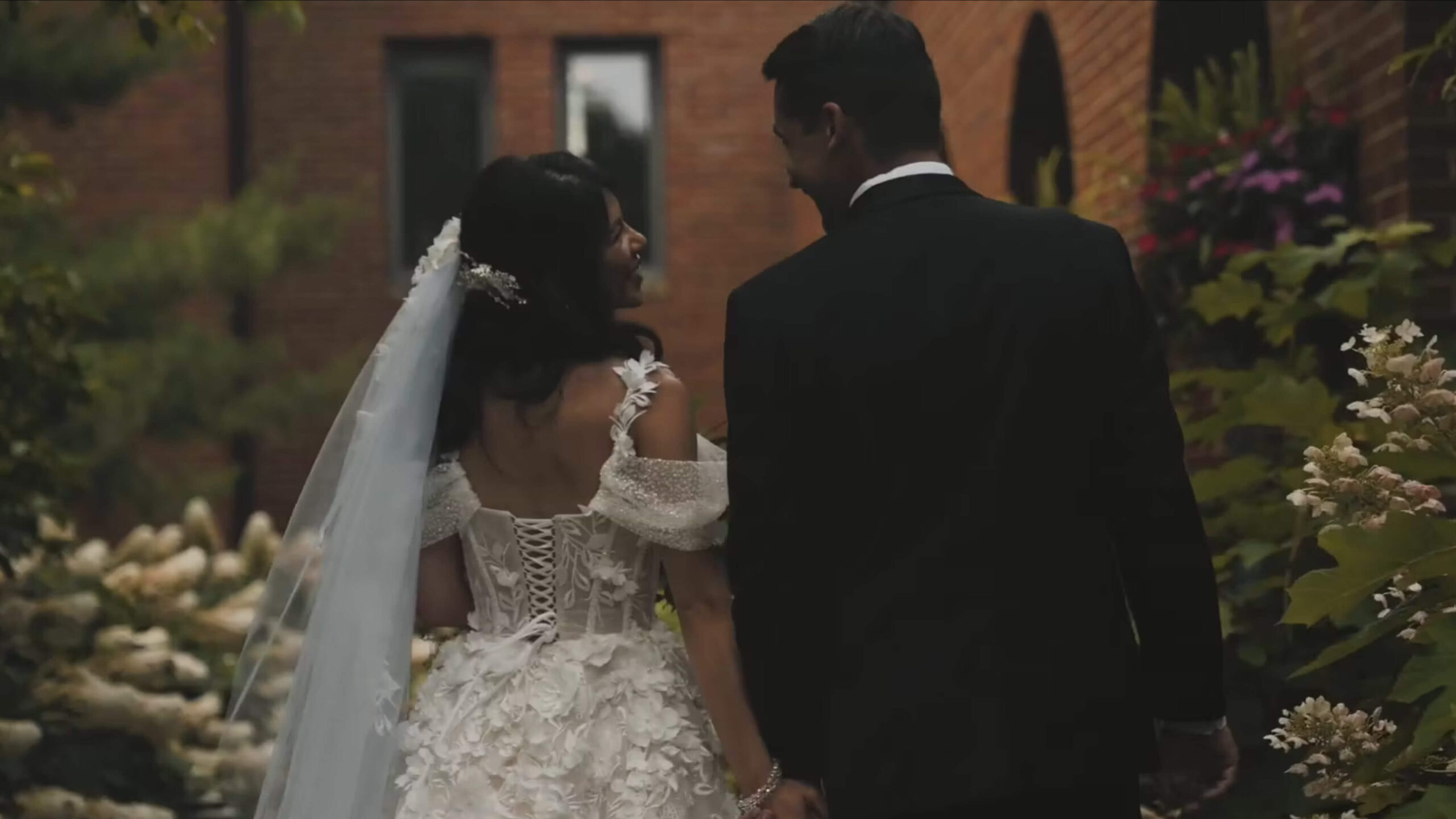 A bride in a white dress and veil walks hand in hand with a groom in a black suit outdoors, in front of a brick building with greenery around them. Both are facing away and looking at each other.