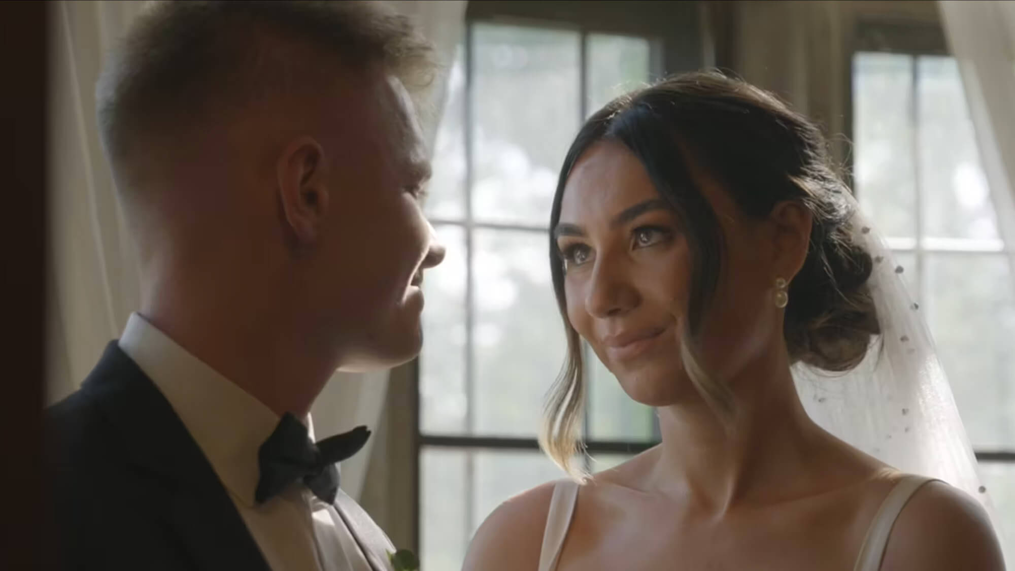 A bride in a white dress and veil looks at a groom in a black tuxedo. They stand indoors near a window with soft daylight illuminating the scene.