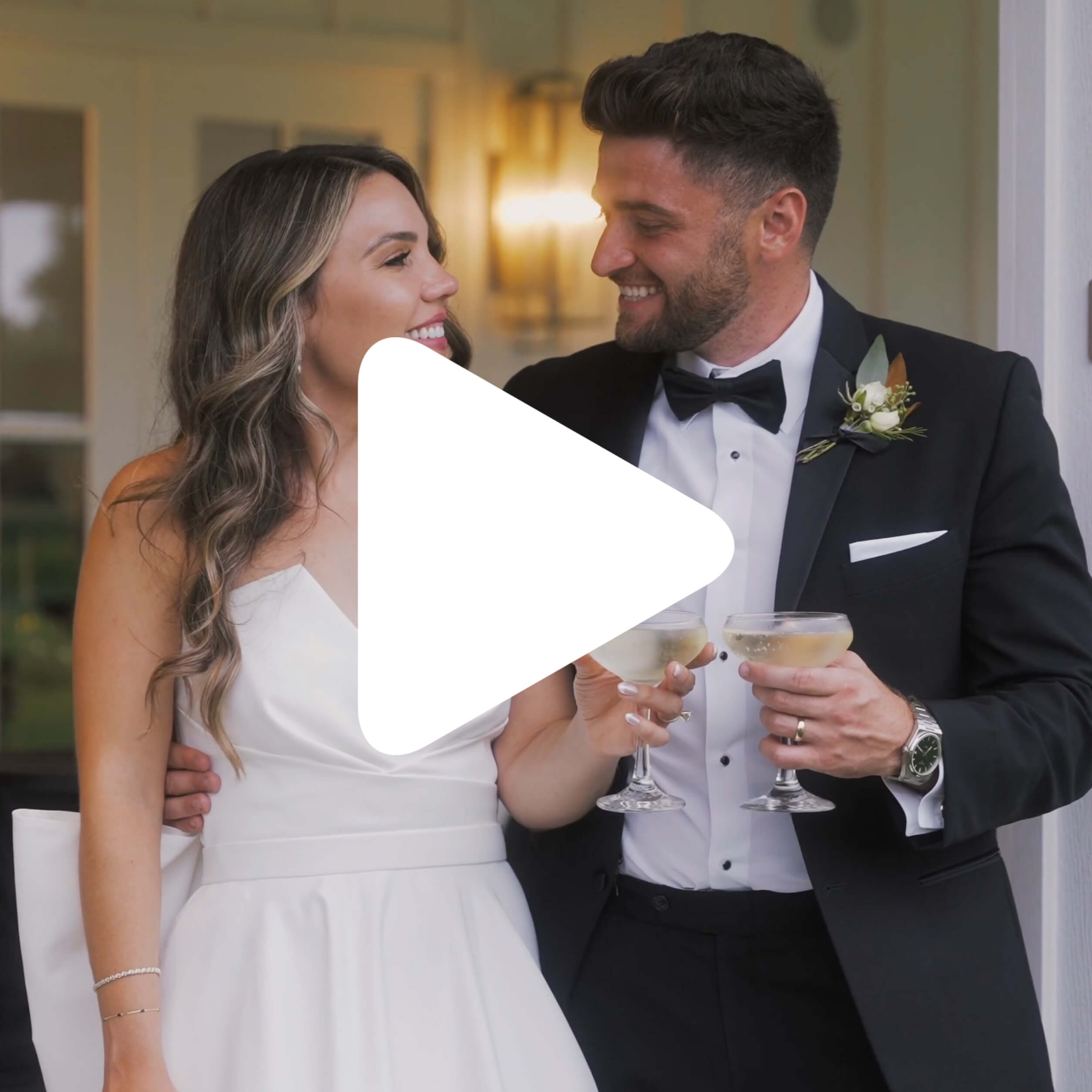 A bride in a white dress and a groom in a black tuxedo stand close together, smiling and holding champagne glasses, with a large play button icon over the center of the image.