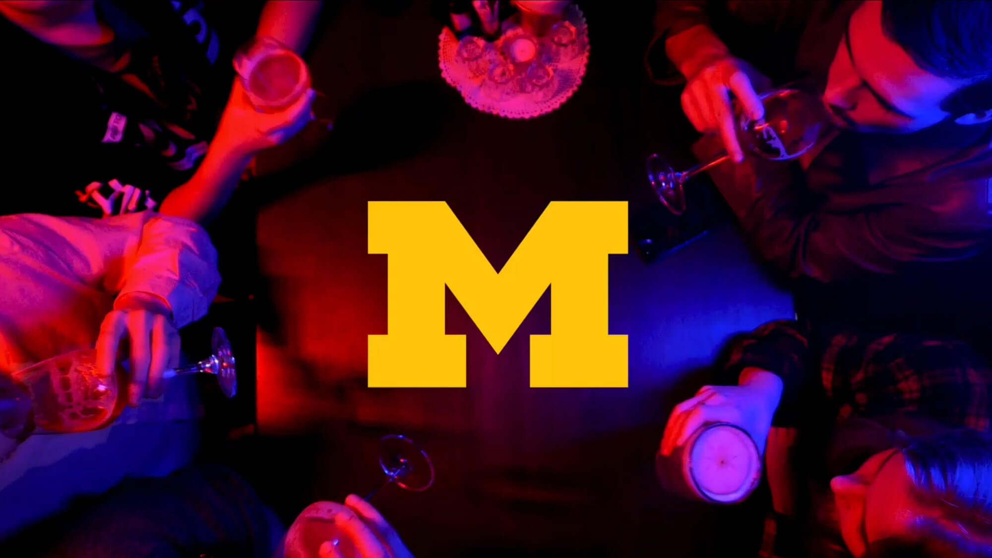 Overhead view of four people holding drinks around a table with a cake, illuminated by red and blue lights. A large yellow letter "M" is centered in the image.
