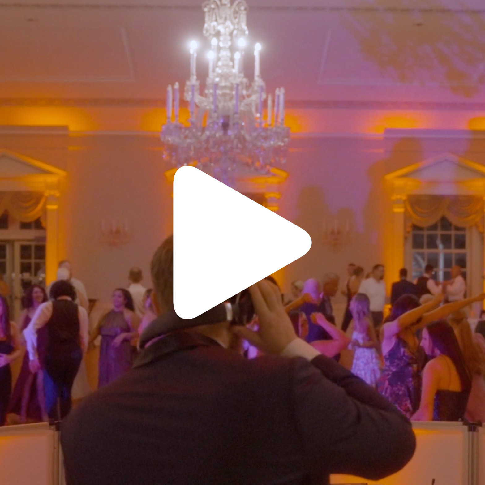 A person facing a crowd at a formal event with a chandelier overhead and purple-orange lighting. A large white play button icon is centered over the image.