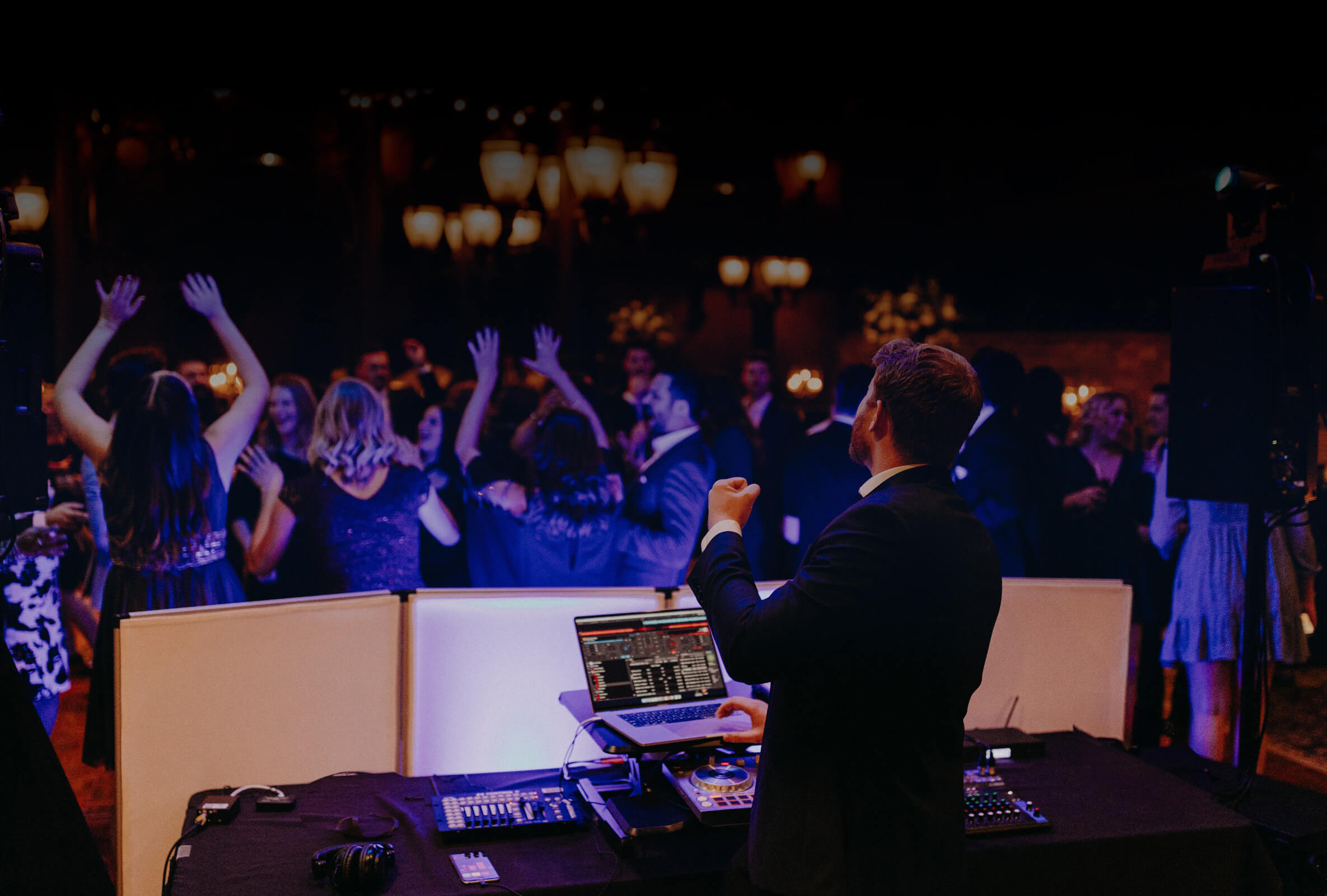 A DJ in a suit plays music on a mixing console while a crowd of people dances and raises their hands at a dimly lit indoor event or party.