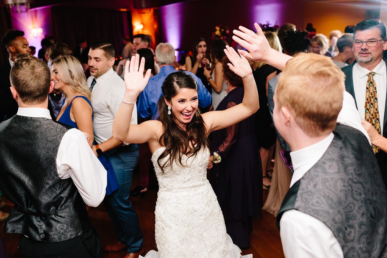 A bride in a white dress smiles with her hands raised while dancing with guests at a lively indoor wedding reception. People around her are dressed formally and appear to be enjoying the celebration.