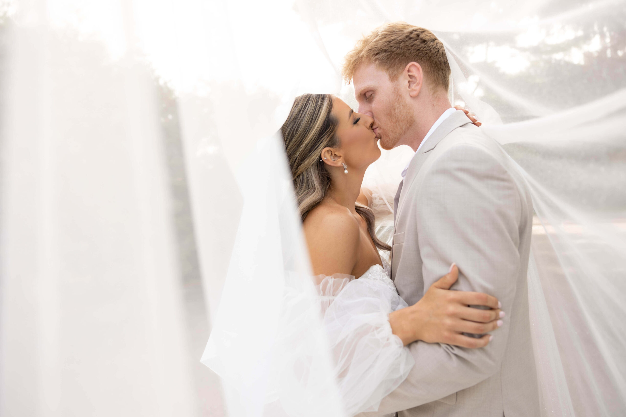 A couple dressed in formal attire shares a kiss while standing close together, partially surrounded by sheer white fabric. The background is softly blurred, and natural light illuminates the scene.