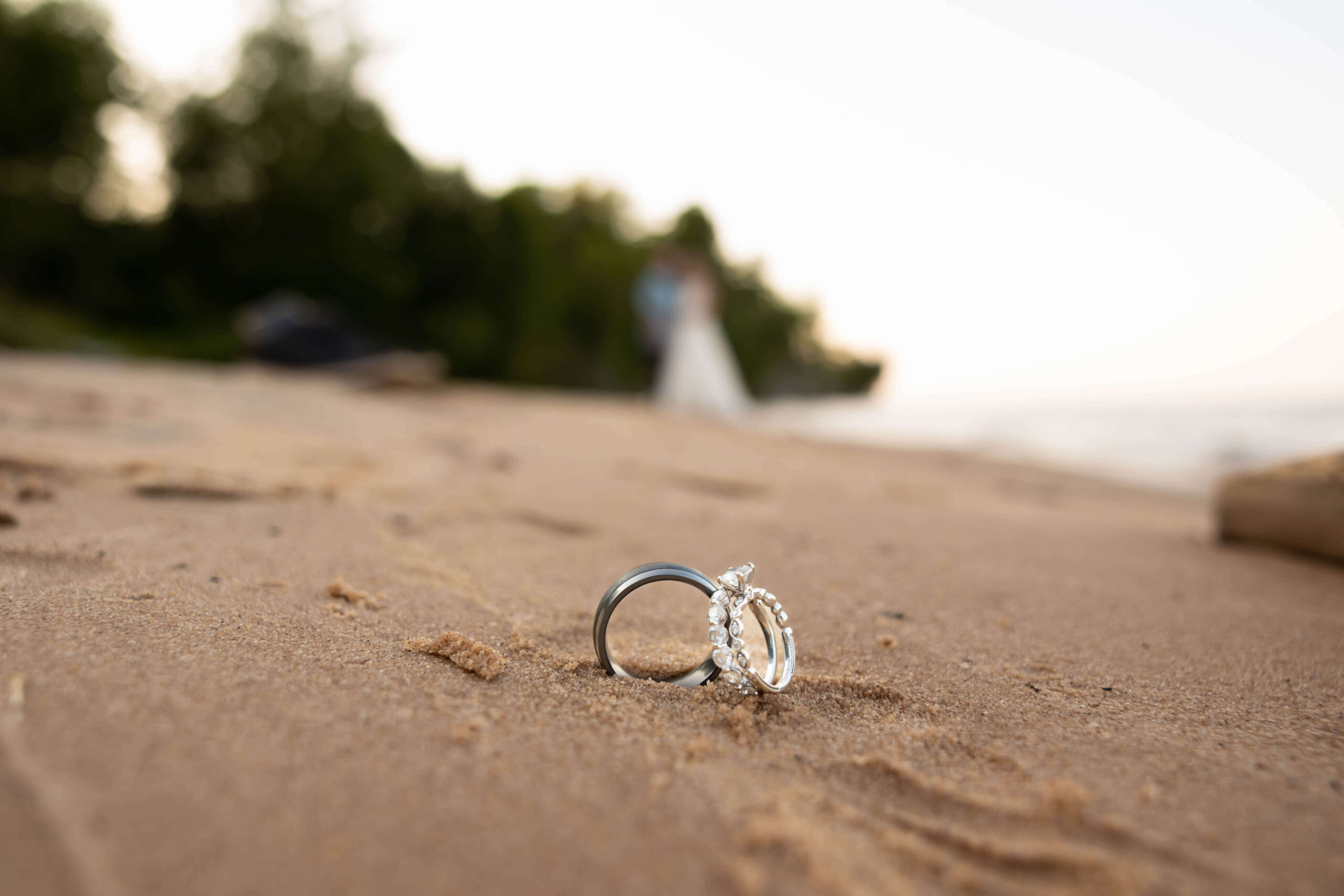 Two rings, one with a diamond and the other a band with small stones, stand upright in the sand on a beach. The background is blurred, showing greenery and two figures near the shoreline.