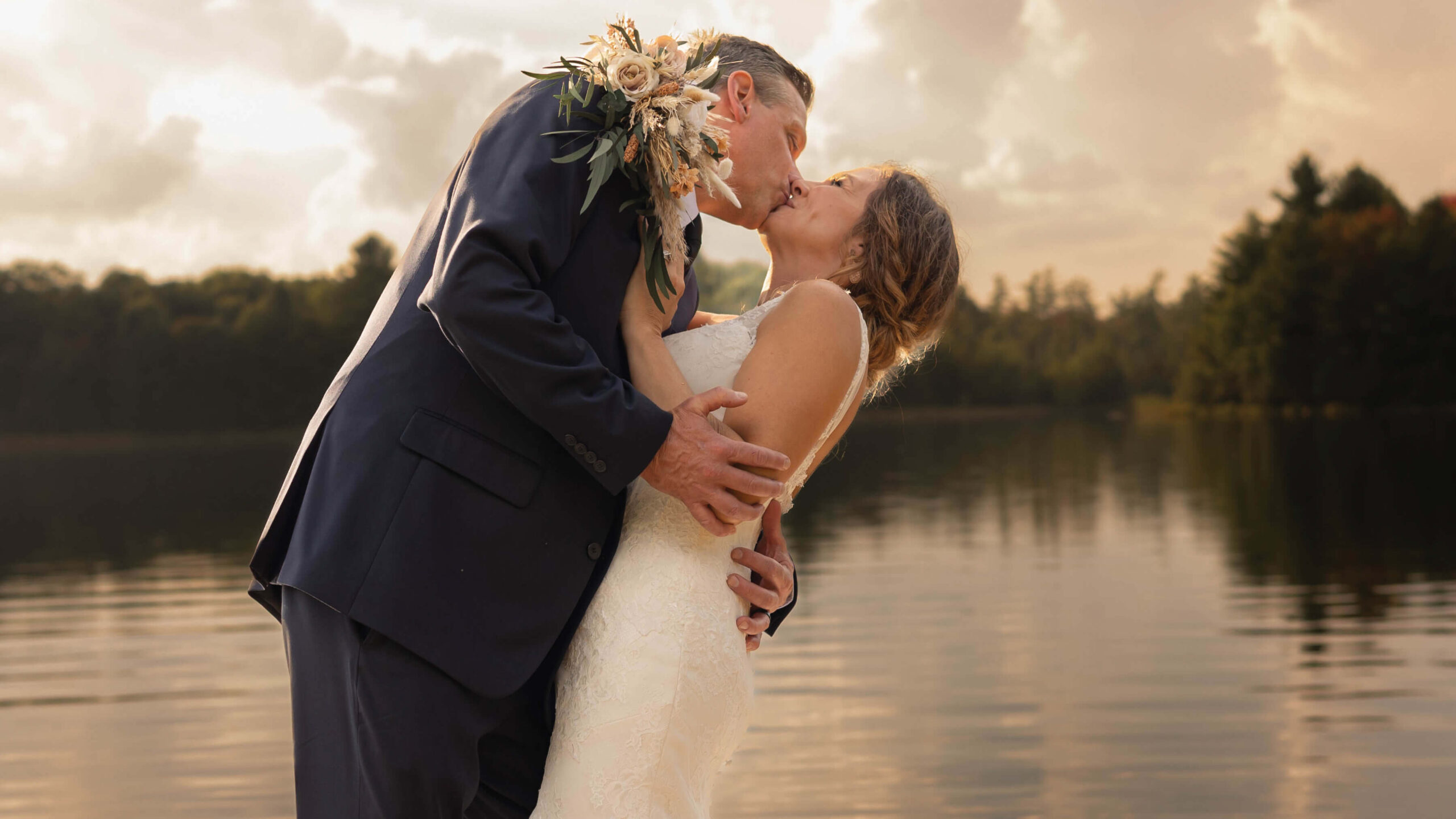 A bride and groom in wedding attire kiss by a lake at sunset. The groom holds a bouquet on his shoulder, and trees are visible in the background under a cloudy sky.