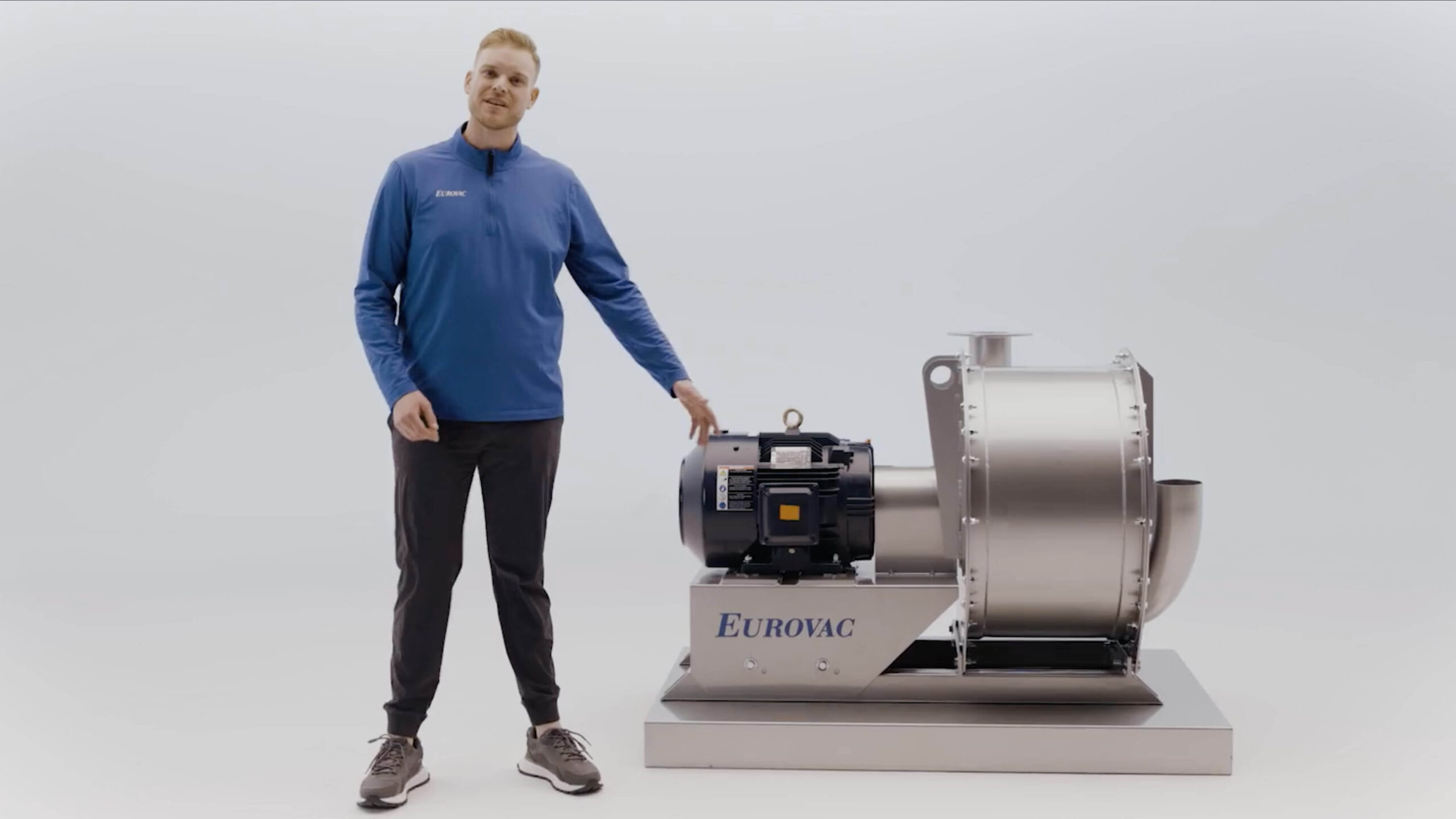 A man in a blue long-sleeve shirt and black pants stands next to a large industrial machine labeled "EUROVAC" in a plain white room, with one hand resting on the machine.