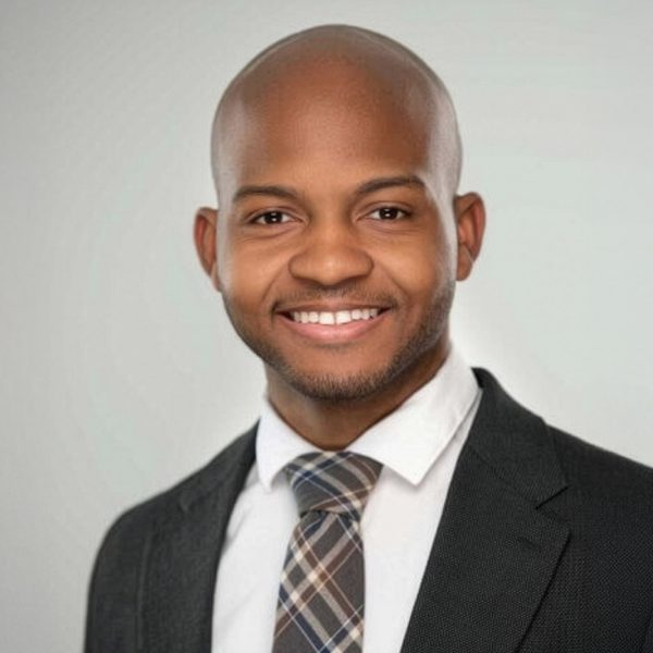 A man wearing a dark suit, white shirt, and plaid tie is smiling at the camera against a plain light gray background.