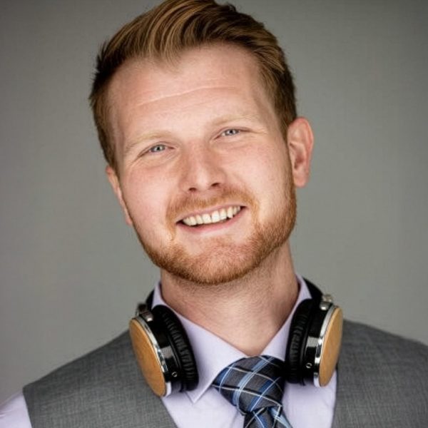 A man with reddish hair and a beard is smiling at the camera. He is wearing a gray vest, patterned tie, light shirt, and has headphones resting around his neck. The background is plain and gray.