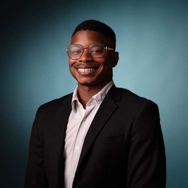 A person wearing glasses, a white shirt, and a black blazer is smiling in front of a solid teal background. The person is facing the camera in a professional portrait style.