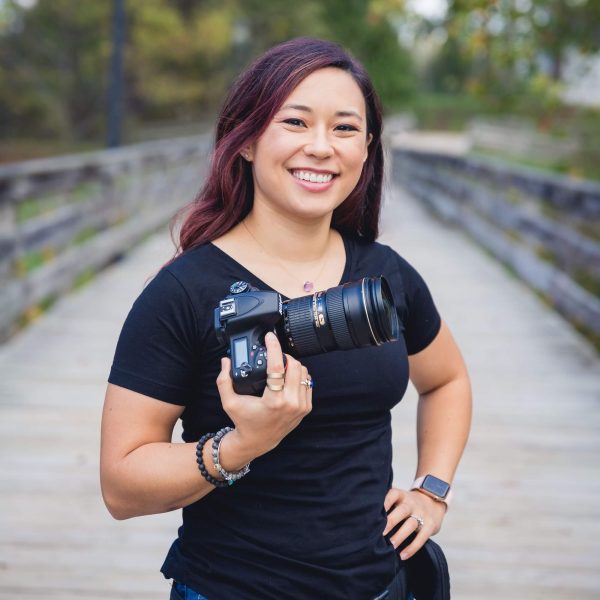 A person with long brown hair, wearing a black t-shirt and jeans, stands outdoors on a wooden bridge holding a DSLR camera and smiling at the camera. Trees and greenery are visible in the background.