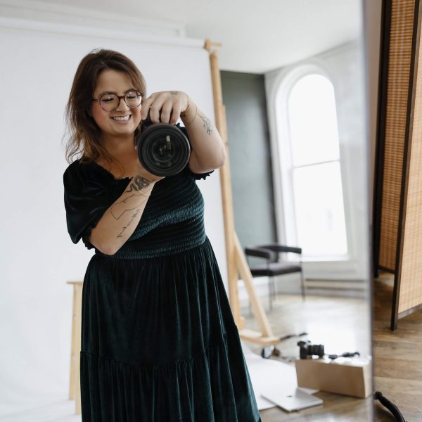 A person with glasses and tattoos is smiling while taking a mirror selfie with a camera in a well-lit room, which contains a tripod, a backdrop, and scattered objects on the floor.