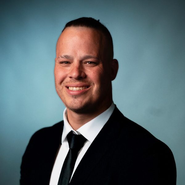 A man in a black suit, white shirt, and black tie is smiling while posing for a formal portrait against a plain blue background.