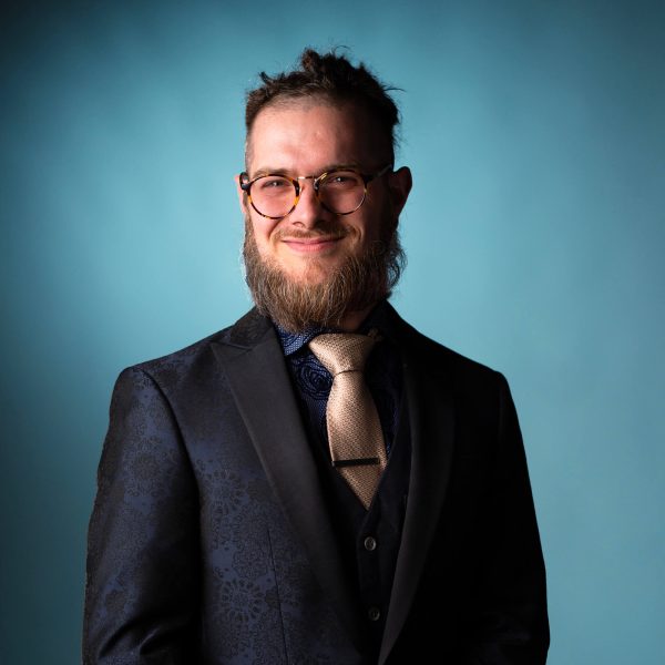 A man with glasses and a beard wears a dark patterned suit, a gold tie, and a dark shirt, standing against a plain blue background and smiling slightly.