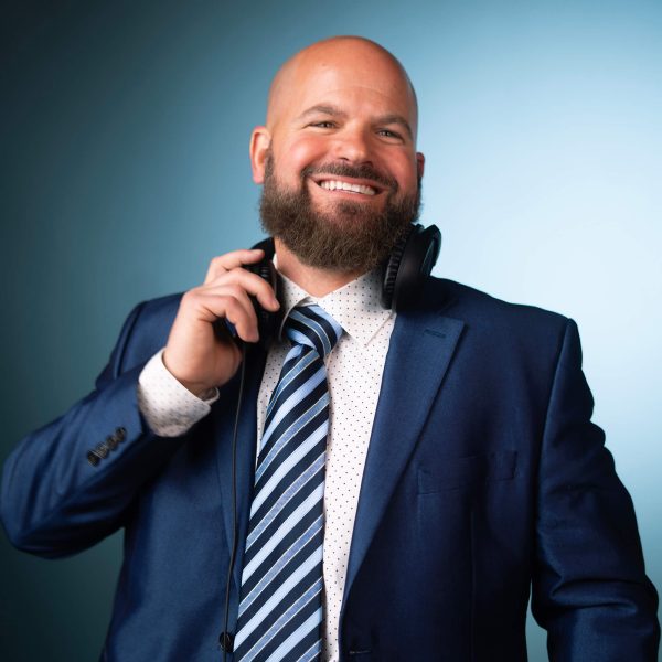 A man with a bald head and a full beard smiles while holding a pair of headphones around his neck. He is wearing a blue suit, a white shirt with small dots, and a striped tie. The background is blue.