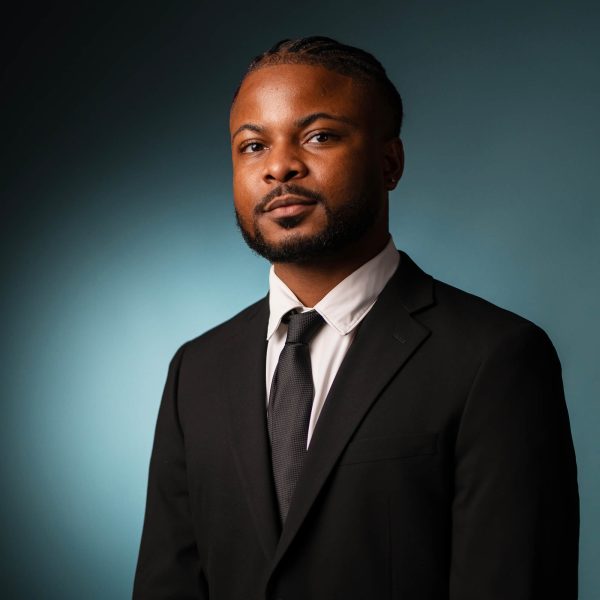 A man wearing a black suit, white dress shirt, and black tie poses against a gradient blue background. He has short braids and a trimmed beard, and he looks confidently at the camera.