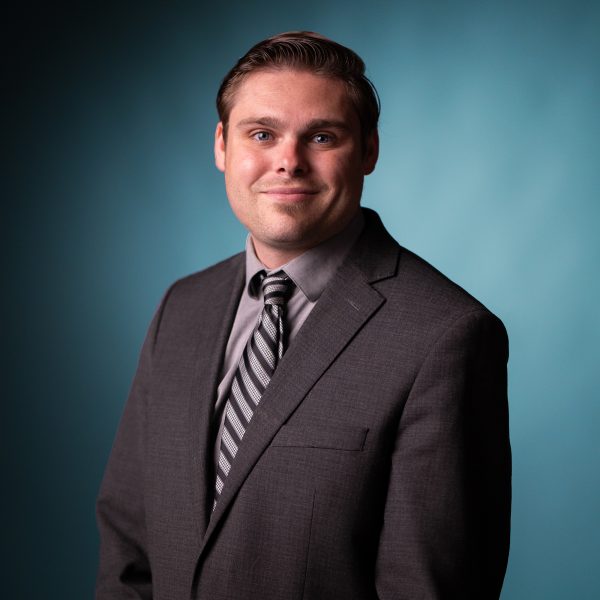 A man with short brown hair wearing a gray suit, striped tie, and light gray shirt poses against a plain blue background. He is facing forward and smiling slightly.