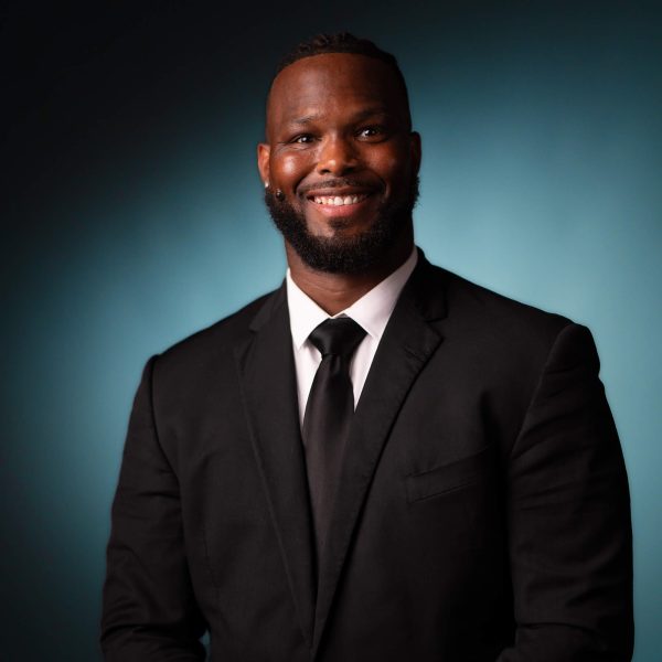 A man wearing a black suit, white shirt, and black tie is smiling at the camera against a blue gradient background.