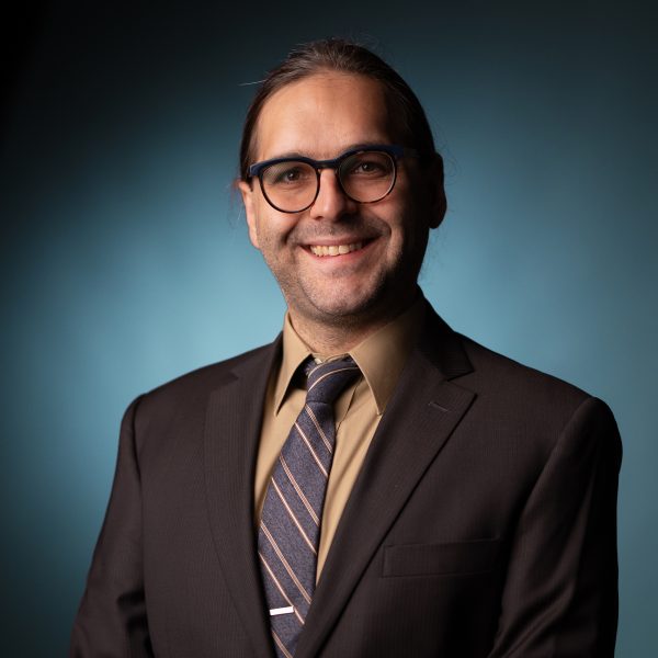 A person wearing glasses, a dark suit, a beige shirt, and a striped tie stands smiling in front of a blue gradient background.