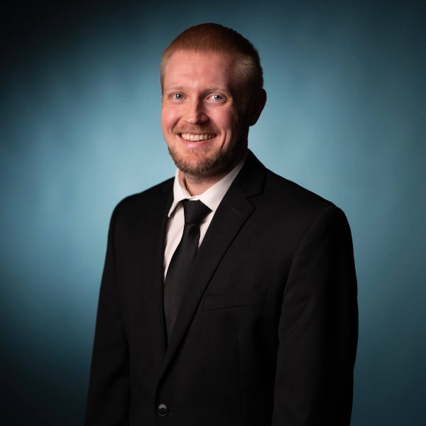 A man with short light hair and a beard is wearing a black suit, white shirt, and black tie. He is standing against a blue gradient background and smiling at the camera.