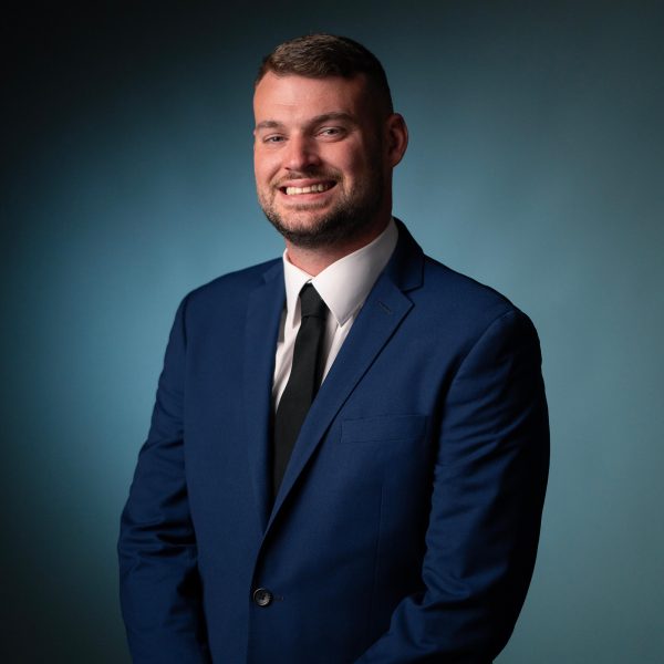 A man with short hair and a trimmed beard is wearing a blue suit, white shirt, and black tie. He is smiling and standing in front of a plain, teal background.
