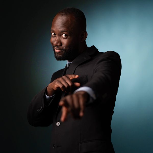 A man in a black suit points toward the camera and smiles, posing against a dark blue gradient background.