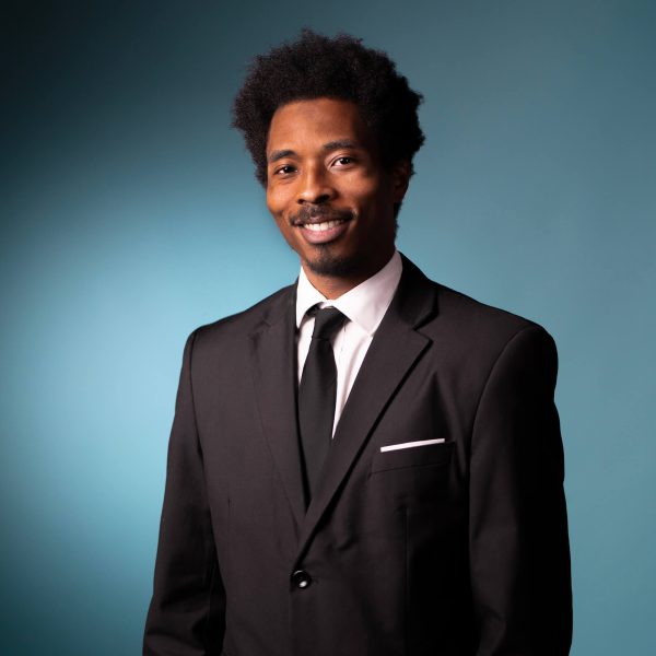 A man wearing a black suit, white dress shirt, and black tie stands in front of a blue background, smiling slightly at the camera.