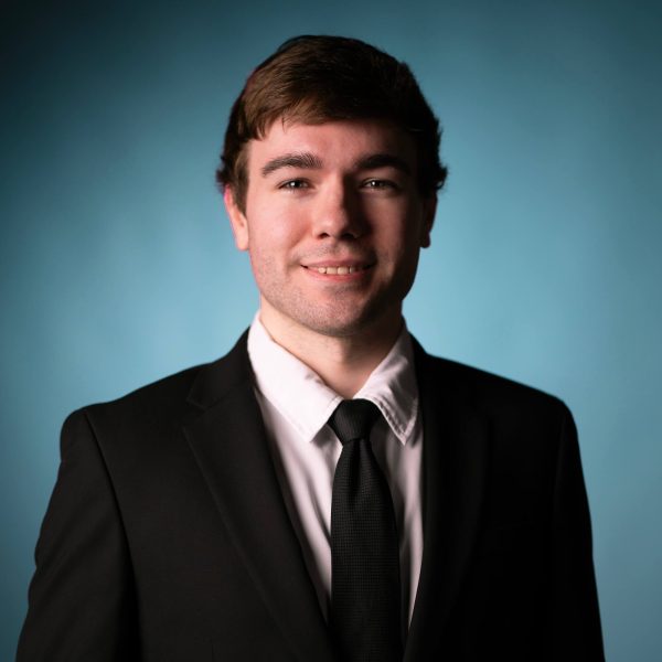 A young man with short brown hair wearing a black suit, white dress shirt, and black tie stands in front of a plain blue background, smiling slightly at the camera.