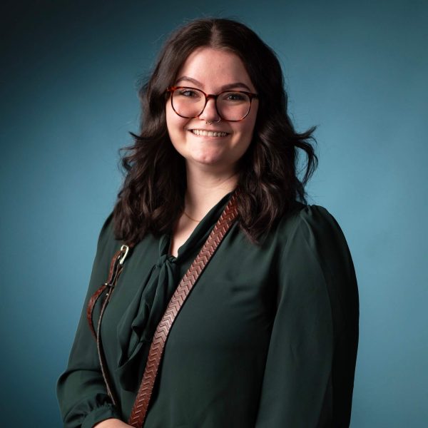 A woman with wavy dark hair and glasses stands in front of a blue background. She is wearing a dark green blouse and a brown strap across her shoulder, smiling at the camera.