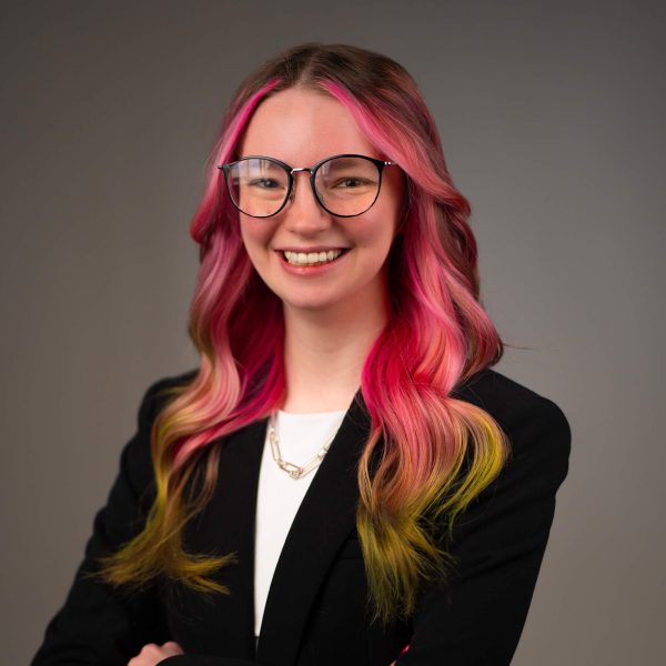 A person with long wavy hair dyed pink with yellow tips is wearing glasses, a black blazer, a white top, and layered necklaces, and is smiling at the camera against a neutral background.