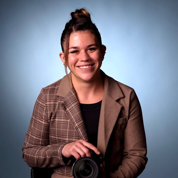 A person with brown hair in a top bun, wearing a plaid blazer and black shirt, sits against a blue background and holds a camera, smiling at the camera.