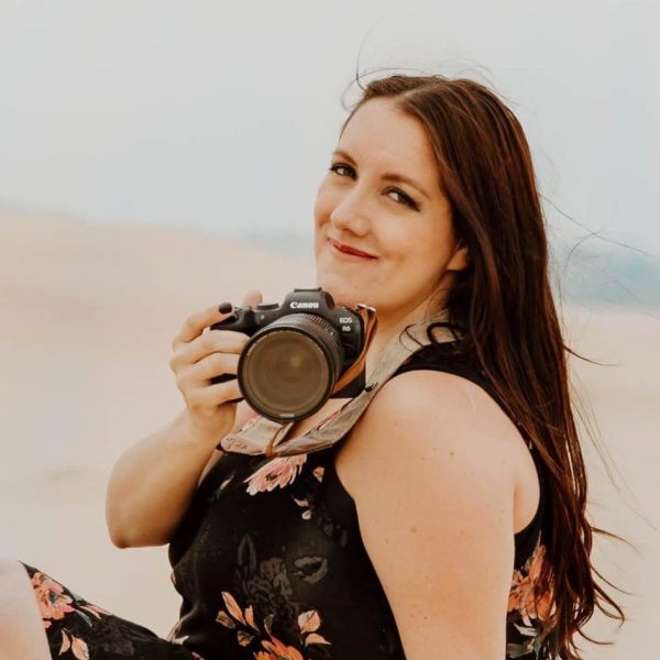 A woman with long brown hair, wearing a black floral dress, holds a Canon camera and smiles at the camera. The background is an outdoor, sandy area with a pale sky.